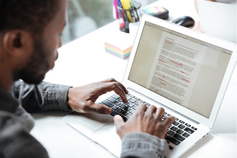 Un homme se tient devant un ordinateur sur lequel on aperçoit un texte dont certains mots sont surlignés en rouge.